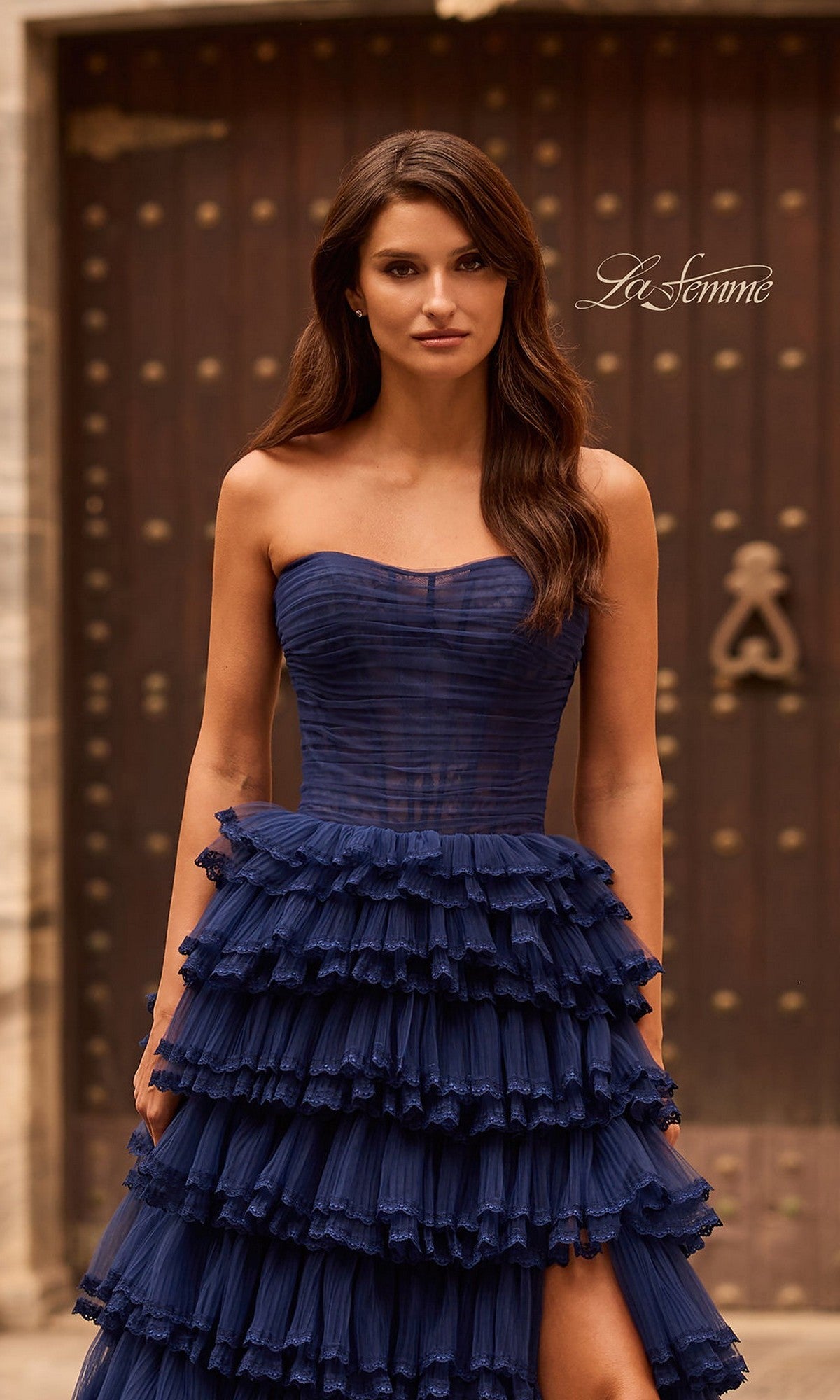 A woman in the La Femme 33727 long prom dress, featuring strapless tiered ruffles in navy blue, stands before a grand wooden door with ornate metal details.